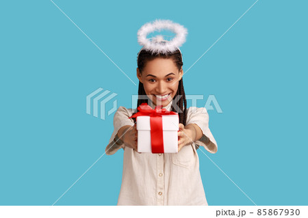 Portrait of cheerful woman with black dreadlocks and halo overhead holding gift box, charity organization, religion, wearing white shirt. Indoor studio shot isolated on blue background. 85867930