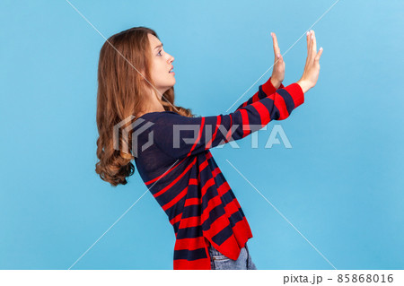 Stop, I am afraid. Side view of scared young woman wearing striped casual style sweater holding hands, looking terrified and desperate. Indoor studio shot isolated on blue background. 85868016