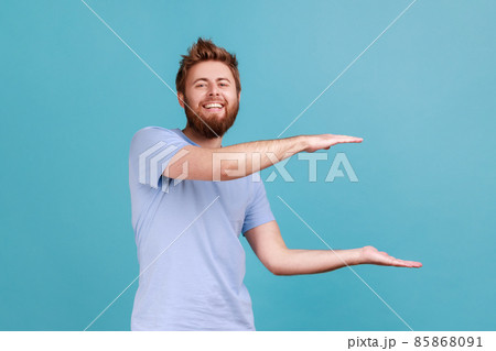 Portrait of young adult positive optimistic bearded man presenting area for advertisement, looking at camera with toothy smile and optimism. Indoor studio shot isolated on blue background. 85868091