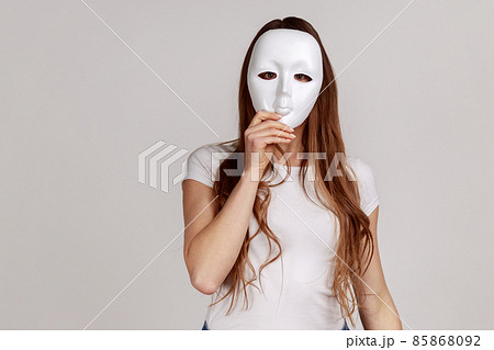 Portrait of anonymous dark haired woman covering her face with white mask, hiding personality, conspiracy and privacy, wearing white T-shirt. Indoor studio shot isolated on gray background. 85868092
