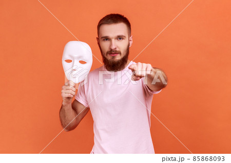 Portrait of serious bearded man pointing finger to camera and holding white mask in his hand, wants to change personality, wearing pink T-shirt. Indoor studio shot isolated on orange background. 85868093