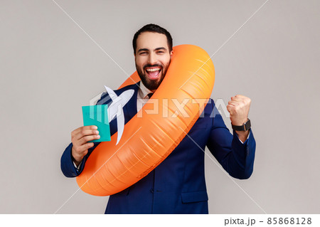 Happy bearded businessman holding orange rubber ring, paper plane and passport, clenching fist, long awaited vacation, wearing official style suit. Indoor studio shot isolated on gray background. 85868128
