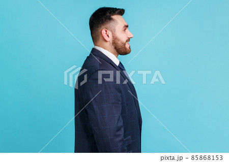 Profile portrait of young adult handsome bearded businessman wearing official style suit looking ahead with toothy smile, optimism, success. Indoor studio shot isolated on blue background. 85868153
