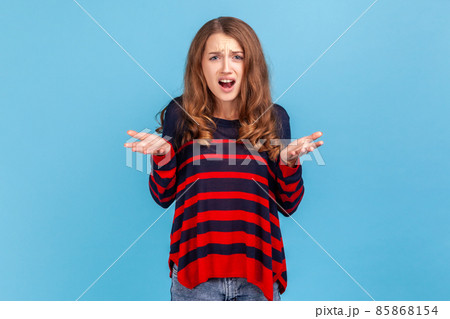 Nervous disappointed woman wearing striped casual style sweater spreading hands and shouting asking what do you want from me. annoyed expression. Indoor studio shot isolated on blue background. 85868154