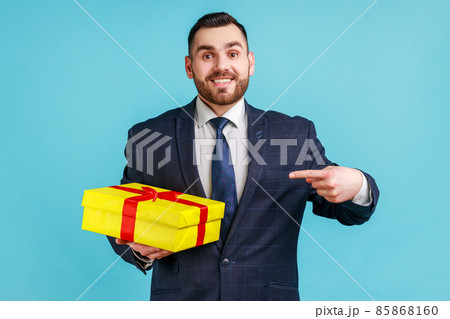 Happy man with beard wearing official style suit standing, holding yellow gift box, pointing finger, looking at camera with toothy smile. Indoor studio shot isolated on blue background. 85868160