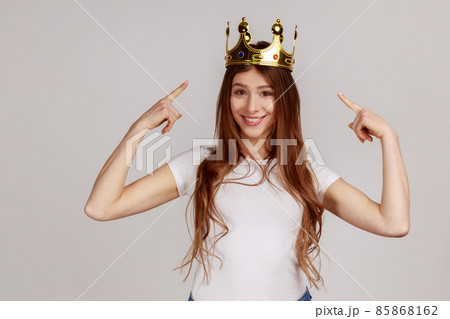 Happy cheerful woman pointing fingers on golden crown on her head, showing her leadership qualities, concept of authority, wearing white T-shirt. Indoor studio shot isolated on gray background. 85868162