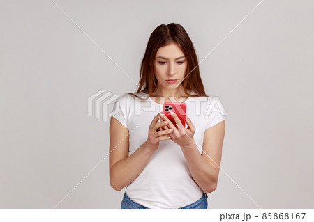 Portrait of concentrated woman using smart phone, typing message or checking social networks, writing post, serious expression, wearing white T-shirt. Indoor studio shot isolated on gray background. Portrait of concentrated woman using smart phone, typing message or checking social networks, writing post, serious expression, wearing white T-shirt. Indoor studio shot isolated on gray background. 85868167