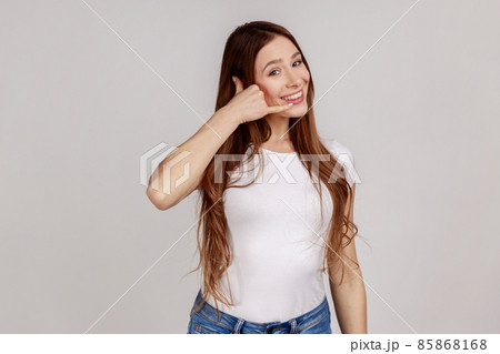Hey handsome, call me. Beautiful woman flirting holding fingers near ear showing call gesture with toothy smile, answering call, wearing white T-shirt. Indoor studio shot isolated on gray background. Hey handsome, call me. Beautiful woman flirting holding fingers near ear showing call gesture with toothy smile, answering call, wearing white T-shirt. Indoor studio shot isolated on gray background. 85868168