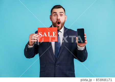 Surprised businessman with beard wearing official style suit holding sale card and smartphone , shocked with amazing online sell-out. Indoor studio shot isolated on blue background. Surprised businessman with beard wearing official style suit holding sale card and smartphone , shocked with amazing online sell-out. Indoor studio shot isolated on blue background. 85868171
