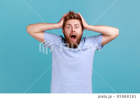 Portrait of pleasant looking cheerful bearded man with mouth open in amazement, expressing shock, astonishment, keeping hands on his head. Indoor studio shot isolated on blue background. Portrait of pleasant looking cheerful bearded man with mouth open in amazement, expressing shock, astonishment, keeping hands on his head. Indoor studio shot isolated on blue background. 85868181