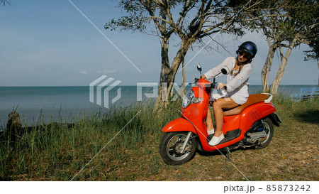 Woman on red motorbike in white clothes drive on forest road trail trip. One girl caucasian tourist go on scooter, nearby tropical palm tree. 85873242