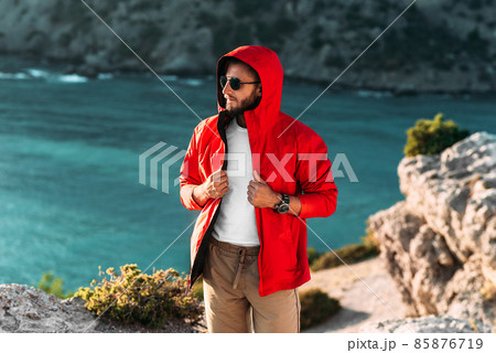 Portrait of a brutal bearded man in a red jacket by the sea. Male portrait on the background of a seascape. A traveler on the background of the sea coast. A man in a jacket at sunset 85876719