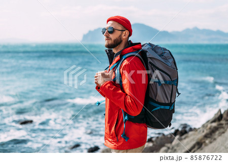 A bearded traveler with a backpack on the background of the sea. Portrait of a traveler in red clothes and sunglasses. A tourist with a backpack stands against the background of the sea. Copy space 85876722
