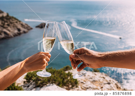 Hands holding champagne glasses over the sea. Romantic vacation. Two hands holding champagne glasses on the background of the sea. A couple in love drinks champagne on the seashore. Copy space Hands holding champagne glasses over the sea. Romantic vacation. Two hands holding champagne glasses on the background of the sea. A couple in love drinks champagne on the seashore. Copy space 85876898