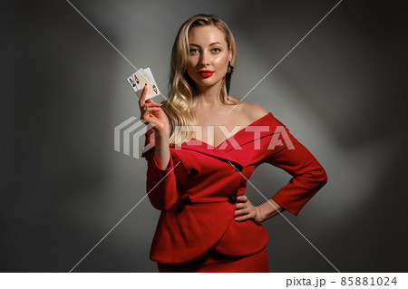 Blonde woman in red dress and black earrings. She is showing two playing cards, posing on gray studio background. Poker, casino. Close-up, copy space Blonde woman in red dress and black earrings. She is showing two playing cards, posing on gray studio background. Poker, casino. Close-up, copy space 85881024