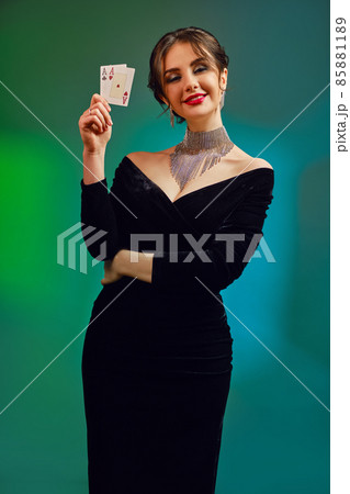 Brunette girl in black dress, necklace and earrings. Smiling, showing two playing cards, posing on colorful background. Poker, casino. Close-up Brunette girl in black dress, necklace and earrings. Smiling, showing two playing cards, posing on colorful background. Poker, casino. Close-up 85881189