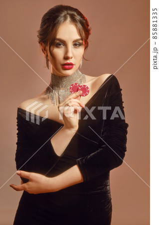 Brunette model in black dress and shiny jewelry. She is showing two red chips, posing against brown studio background. Poker, casino. Close-up Brunette model in black dress and shiny jewelry. She is showing two red chips, posing against brown studio background. Poker, casino. Close-up 85881335