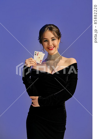 Brunette woman with bare shoulders, in black dress and shiny jewelry. Smiling, showing two aces, posing on purple background. Poker, casino. Close-up Brunette woman with bare shoulders, in black dress and shiny jewelry. Smiling, showing two aces, posing on purple background. Poker, casino. Close-up 85882220