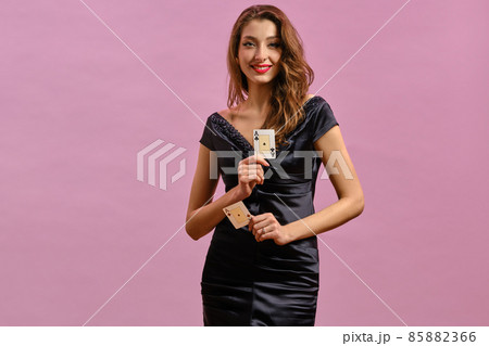 Brunette lady with earring in nose, in black dress. Showing two playing cards, smiling, posing on pink studio background. Poker, casino. Close-up 85882366