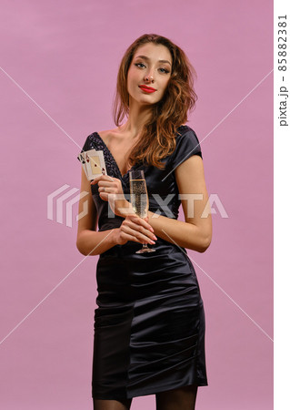 Brunette lady with earring in nose, in black dress. Holding glass of champagne, two aces, smiling, posing on pink background. Poker, casino. Close-up Brunette lady with earring in nose, in black dress. Holding glass of champagne, two aces, smiling, posing on pink background. Poker, casino. Close-up 85882381
