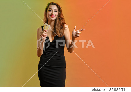 Girl in black dress. Holding two playing cards, smiling, pointing at something, posing colorful background. Poker, casino. Template, mockup. Close up 85882514