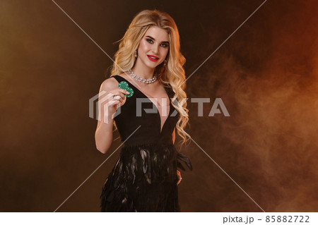 Blonde female in black dress with feathers and necklace. Smiling, showing two green chips, posing on dark smoky background. Poker, casino. Close-up Blonde female in black dress with feathers and necklace. Smiling, showing two green chips, posing on dark smoky background. Poker, casino. Close-up 85882722