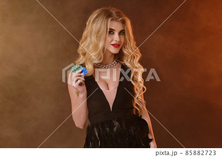 Blonde woman in black dress and necklace. Smiling, showing green and blue chips, posing on dark smoky studio background. Poker, casino. Close-up Blonde woman in black dress and necklace. Smiling, showing green and blue chips, posing on dark smoky studio background. Poker, casino. Close-up 85882723