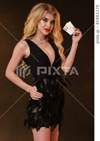 Blonde female in black dress and necklace. She is smiling, showing two aces, posing sideways on black smoky background. Poker, casino. Close-up 85883270