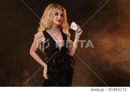 Blonde model in black dress with feathers and necklace. Smiling, showing two playing cards, posing on black smoky background. Poker, casino. Close-up 85883272
