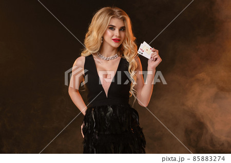 Blonde lady in black dress with feathers and necklace. She is smiling, showing two aces, posing on black smoky background. Poker, casino. Close-up 85883274