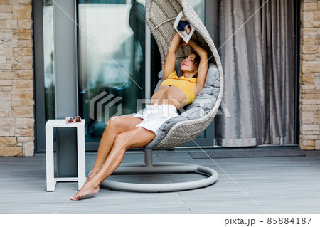 A young woman is resting in a suspended swing on the terrace of the house with a magazine in her hands A young woman is resting in a suspended swing on the terrace of the house with a magazine in her hands 85884187