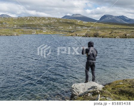 Fisherman, angler figure at northern landscape, tundra in Swedish Lapland with blue artic lake, green hills and mountains. Fishing at Padjelantaleden hiking trail. Summer day, blue sky, white clouds 85885438