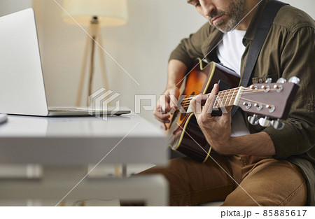 Musical performance online. Man plays acoustic guitar at home in front of online audience or provides music lesson using laptop. Cropped image of serious man with guitar in his hands. Selective focus. 85885617