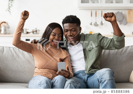 Happy black lovers using smartphone together and gesturing, home interior Happy black lovers using smartphone together and gesturing, home interior 85888386