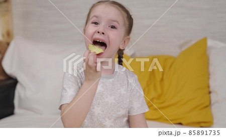 little girl eating golden potato chips sitting on the bed in the bedroom, fast food for kid, unhealthy baby food, enjoying the taste of potato spices, girl's favorite snack at home, happy daughter little girl eating golden potato chips sitting on the bed in the bedroom, fast food for kid, unhealthy baby food, enjoying the taste of potato spices, girl's favorite snack at home, happy daughter 85891735