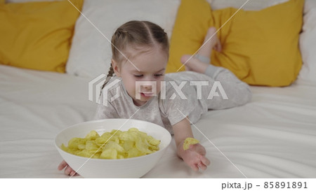 little child eats crispy fries lying on the bed, kid bed food, delicious baby food chips, snacks, happy family, children's life at home, baby girl gnaws fried vegetable, not healthy human food. little child eats crispy fries lying on the bed, kid bed food, delicious baby food chips, snacks, happy family, children's life at home, baby girl gnaws fried vegetable, not healthy human food. 85891891