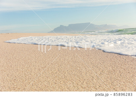 Sea wave foam on shore at sunny beach with mountains in distant background 85896283