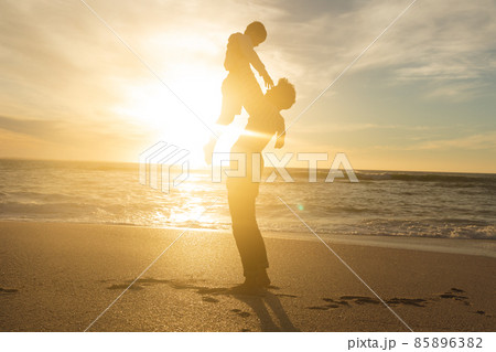 Side view of back lit biracial father holding aloft son on shore at beach against sky during sunset Side view of back lit biracial father holding aloft son on shore at beach against sky during sunset 85896382