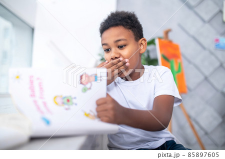 Boy looking at book shyly covering mouth with hand Boy looking at book shyly covering mouth with hand 85897605