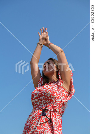 Relaxing young millennial woman in red dress against blue sky. Beautiful smiling girl on summer having fun. Enjoying sun 85902858
