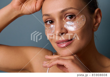 Close-up portrait of an attractive smiling woman applying gold medical hydrogel collagen eye patches to reduce bags under her eyes and puffiness, posing at camera against a gray background. Copy space 85908608