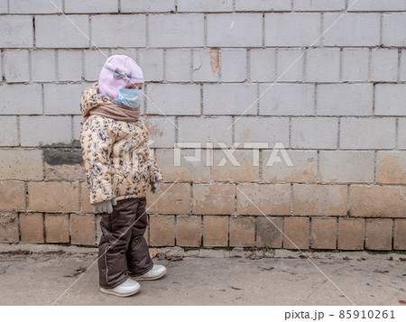 A little child girl in a protective mask on her face stands near an old wall.  85910261