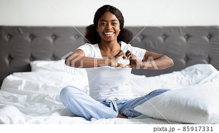 Positive black woman in pajamas sitting on bed, showing heart 85917331