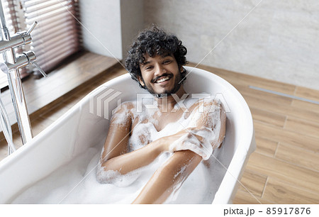 Happy naked indian man taking bath at home, laying in white bathtub with foam and smiling at camera, copy space 85917876