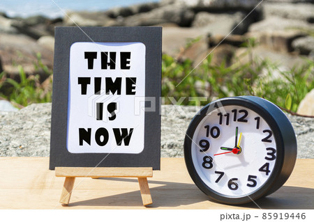 Motivational quote on standing notepad with black alarm clock. The time is now 85919446