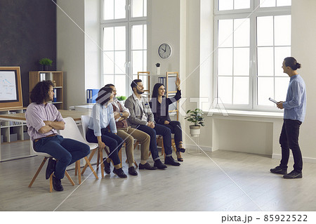 Leader of the startup team listens to the suggestions of his colleagues at a meeting in the office. Woman surrounded by her colleagues sitting on chairs raises her hand wanting to talk about new ideas 85922522
