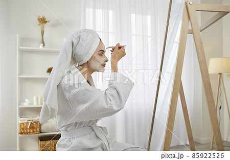 Beautiful young woman in morning after shower with brush applies moisturizing cosmetic mask on her face. Woman in white bathrobe and with towel on her head sitting in front of large mirror in room. 85922526