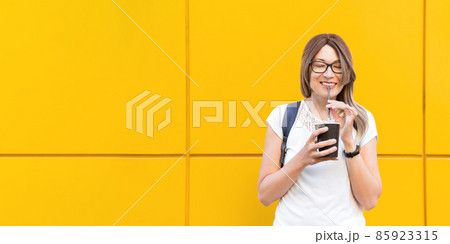 Wide smiling woman drinks coffee with black straw. Portrait of happy female with tasty cappuccino on monochrome yellow wall background. Bright and colorful backdrop. Student with coffee to go. Wide smiling woman drinks coffee with black straw. Portrait of happy female with tasty cappuccino on monochrome yellow wall background. Bright and colorful backdrop. Student with coffee to go. 85923315