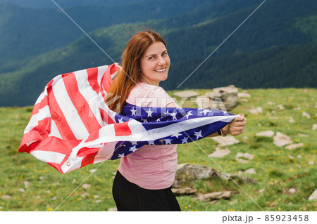 Girl with American Flag looking out at landscape. Young woman stands in the mountains, holding the US flag in her arms high. Flag fluttering in the wind Girl with American Flag looking out at landscape. Young woman stands in the mountains, holding the US flag in her arms high. Flag fluttering in the wind 85923458