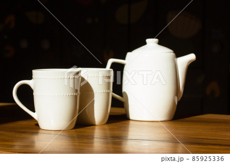 Two white ceramic or porcelain tea cups and tea or coffee kettle pot stand on a wooden table at sunny morning in a kitchen. White kitchen dishware. White ware on dark black background. Selective focus Two white ceramic or porcelain tea cups and tea or coffee kettle pot stand on a wooden table at sunny morning in a kitchen. White kitchen dishware. White ware on dark black background. Selective focus 85925336